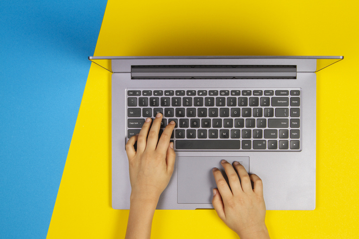 Kid hands typing on laptop computer keyboard, top view, yellow and blue background